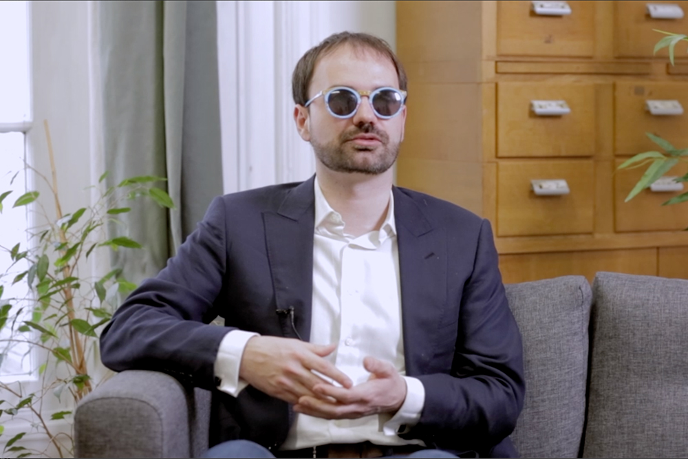 Photo de Thibaut de Martimprey, assis sur un canapé gris. Il porte une chemise blanche et un costume gris foncé, ainsi que des lunettes noires à monture bleu ciel.