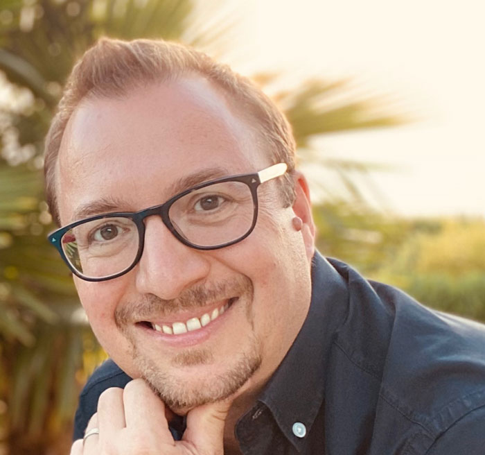Photo de David Geiswiller, de face. Il porte des lunettes à monture noire et une chemise bleue foncé ; il sourit et se tient le menton avec sa main gauche. Il a des cheveux courts, blond-châtain, ainsi qu'une fine moustache et un bouc.